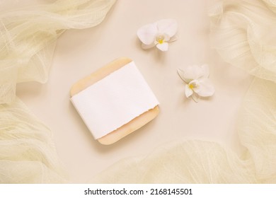 Hanmade soap bar near white orchid flowers and tulle on light yellow, top view, mockup. Skincare beauty product. Exotic natural cosmetics, pastel minimal composition