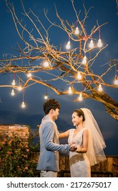 Hanoi City, Vietnam - December 2021: The Bride And Groom Are Happily Together In The Rooftop Garden Under The Romantic Shimmering Lights. The Photo Was Taken At Night.