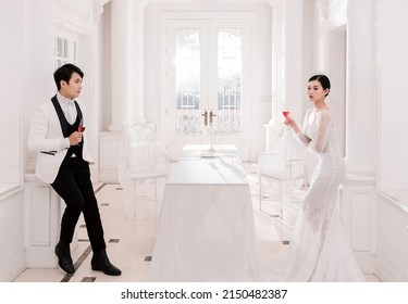 Hanoi City, Vietnam - March 2021: The Elegant Bride And Groom Raise A Glass Of Wine In A Luxurious White Furnished Apartment. Rich And Chic Asian Couple.