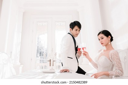 Hanoi City, Vietnam - March 2021: The Elegant Bride And Groom Raise A Glass Of Wine In A Luxurious White Furnished Apartment. Rich And Chic Asian Couple.