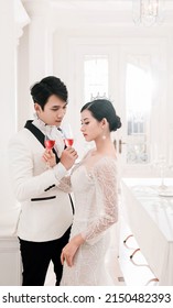 Hanoi City, Vietnam - March 2021: The Elegant Bride And Groom Raise A Glass Of Wine In A Luxurious White Furnished Apartment. Rich And Chic Asian Couple.