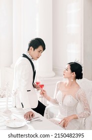 Hanoi City, Vietnam - March 2021: The Elegant Bride And Groom Raise A Glass Of Wine In A Luxurious White Furnished Apartment. Rich And Chic Asian Couple.