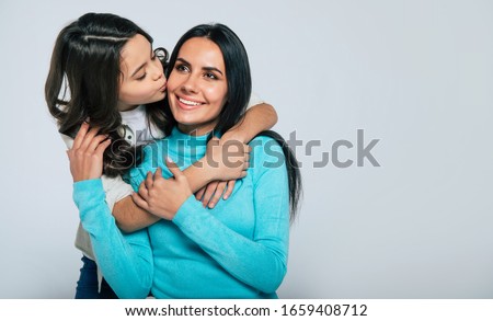 The happiest mother. Young gorgeous mother in blue turtleneck sweater is enjoying the moment when her little daughter is hugging her from the back and kissing in the cheek.