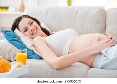 The happiest time for every woman. Beautiful pregnant woman looking at camera and smiling while lying on a couch