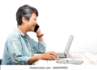 Happily Active Asian Senior Businesswoman With Grey Hair Working On A Laptop While Talking A Cellphone In The Office. Isolated White Background. Active Elderly Concept.