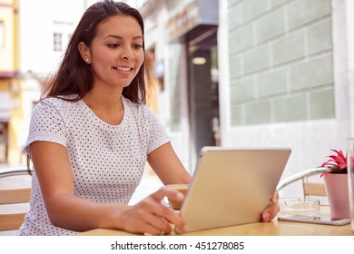 Happily smiling young girl looking at her tablet in a street cafe, relaxing while wearing her hair loose and casual clothing