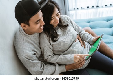 happiness of couples holding the wife's pregnant stomach while using digital tablet, sitting on the couch beside the ornamental plants