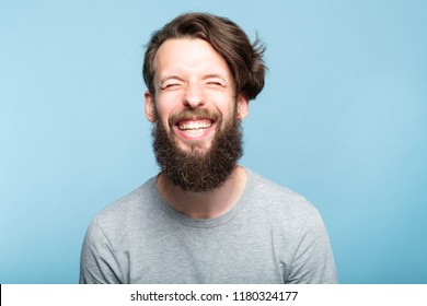 Happiness Enjoyment And Laugh. Exhilarated Man With A Wide Grin. Portrait Of A Young Bearded Hipster Guy On Blue Background. Emotion Facial Expression. Feelings And People Reaction.