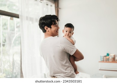 Happiness of fatherhood. Young Asian dad with adorable baby on his hands standing near window at home. Loving father spending time with infant child. Fathers day concept. - Powered by Shutterstock