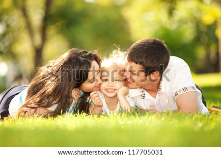 Happiness and harmony in family life. Happy family concept. Young mother and father kissing their daughter in the park. Happy family resting together on the green grass. Family having fun outdoor
