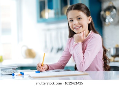 Happiness inside me. Beautiful alert dark-eyed dark-haired girl smiling and writing in her notebook while sitting at the computer and holding her head with her hand - Powered by Shutterstock