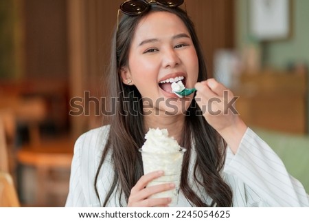 happiness joyful asian young adult female woman teasing yummy smiling hand hold whipped cream in plastic cup,enjoy eating asia woman toothy smile while take a break with glass of dessert in cafe