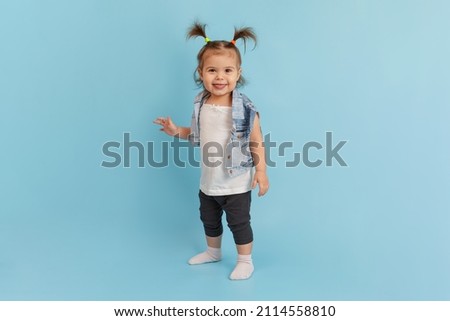 Happiness little girl with two ponytails wearing trendy denim jacket clothes stands in full growth. Happy childhood and trendy children wear concept. Child fashion. Isolated on blue background.