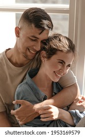 Happiness, love, feeling concept. Pretty young couple smiling, hugging each other and looking down in sunlit room - Shutterstock ID 1953169579
