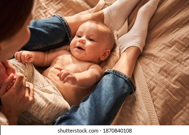 Happiness of motherhood. Back view of the young mom sitting with her baby on bed, looking at son with love and care while playing and holding his legs