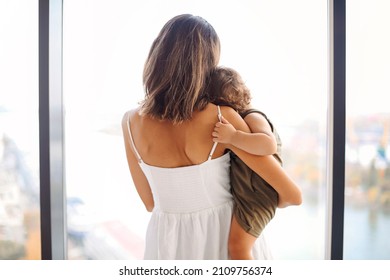 Happiness of motherhood. Rear view of mother wearing white dress with baby looking out window. Mom cuddling her little son infant while spending time at home, enjoying parenting - Powered by Shutterstock