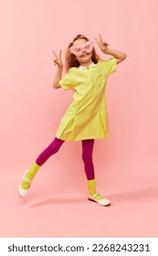 Happiness and positivity. Little cute girl, child with curly hair posing in yellow dress and pink fur glasses on pink studio background. Childhood, emotions, fun, fashion, lifestyle, facial expression