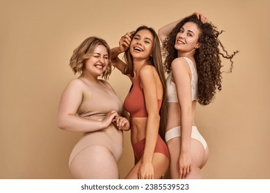 Happiness and self love. Group of three stunning women with beaming smile on faces and different body size posing together in tender lingerie in studio.                            