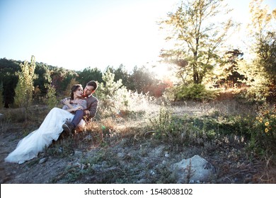 Happiness wedding couple wearing sweater, dress and suit, resting in the nature. The groom hug the bride with love against the beautiful vie of the sun and grass.