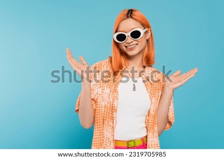 happiness, young asian woman with dyed hair standing in casual attire and sunglasses, gesturing with hands on vibrant blue background, orange shirt, necklace, generation z, red hair