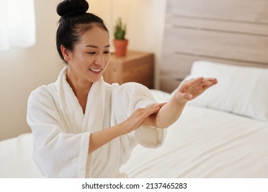 Happly young Asian woman applying body oil on her arms after taking bath at home in the evening