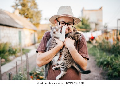 Happpy young bearded farmer holding two little kitten in hands outdoor in village with abstract background. Smiling man in glasses and straw hat playing with funny cute pets. Have fun in countryside. - Powered by Shutterstock