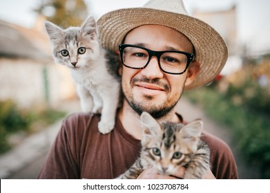 Happpy young bearded farmer holding two little kitten in hands outdoor in village with abstract background. Smiling man in glasses and straw hat playing with funny cute pets. Have fun in countryside.