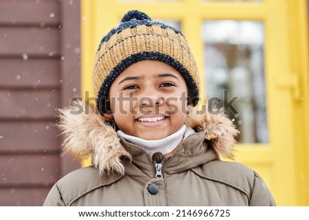 Happu adorable boy in warm winter jacket and striped knitted beanie hat looking at camera while standing against yellow door of country house