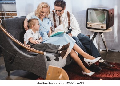 Happy 1950s Style Family Sitting On Sofa And Reading Book Together