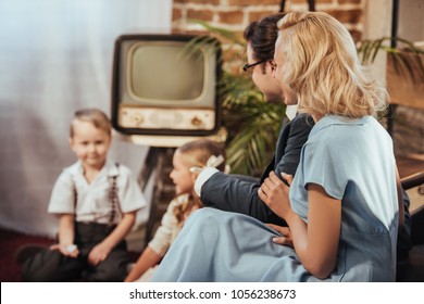 happy 1950s style parents sitting on sofa and lookign at cute little children playing at home