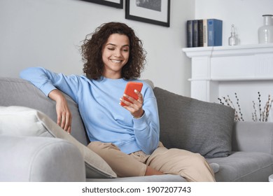 Happy 20s young woman watching social media videos looking at smartphone relaxing on couch, smiling hispanic girl enjoying using online mobile apps doing online shopping on cell phone at home.