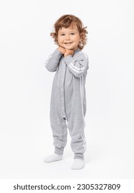 A happy 2-year-old toddler with curly hair in a gray jumpsuit and socks stands and smiles with his hands folded under his chin on a white background. - Shutterstock ID 2305327809