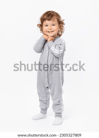 A happy 2-year-old toddler with curly hair in a gray jumpsuit and socks stands and smiles with his hands folded under his chin on a white background.