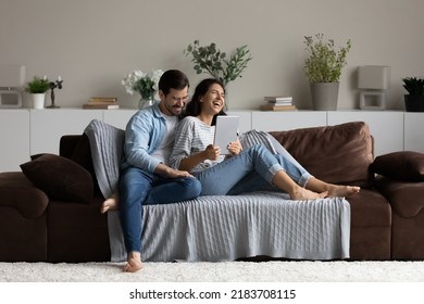 Happy 30s couple relaxing on soft comfortable couch, enjoying leisure time and entertainment in cozy home interior, using tablet, reading book, watching movie, laughing and hugging. Wide shot