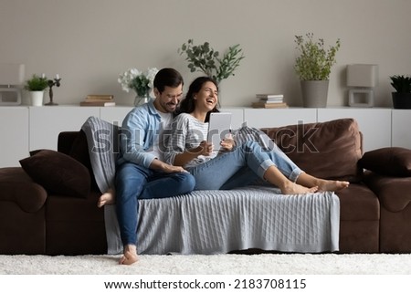 Happy 30s couple relaxing on soft comfortable couch, enjoying leisure time and entertainment in cozy home interior, using tablet, reading book, watching movie, laughing and hugging. Wide shot