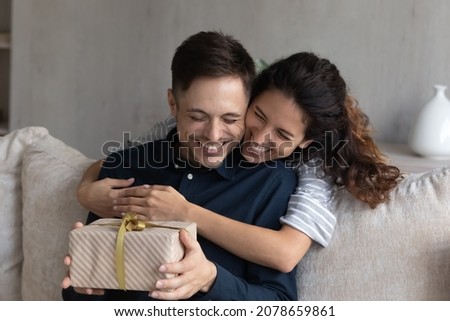 Happy 30s Hispanic wife congratulates husband on birthday, woman makes surprise, gives to beloved man gift box with present, family celebrating life events, hugging expressing love. Congrats concept