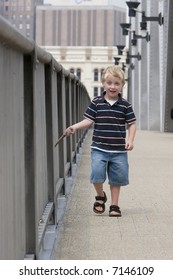 A Happy 4 Year Old Boy Walking Along A Bridge And Striking The Bridge Railing With A Stick