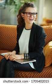 Happy 40 Years Old Woman In White Blouse And Black Jacket With Closed Laptop Sitting On Couch Looking Aside At Modern Home In Sunny Day.