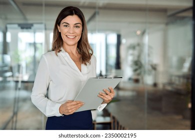 Happy 45 years old Latin professional middle aged business woman corporate leader, happy mature female executive, lady manager standing in office holding clipboard looking at camera, portrait.