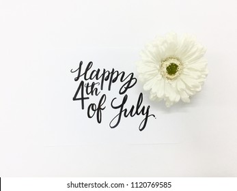 Happy 4th of July hand-lettered on a piece of white paper against a white background with a white daisy on the right side of it flatlay