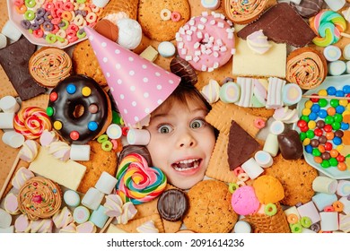 Happy 5 years old girl wearing birthday hat lying under plenty of sweet foods. Kids face surrounded by unhealthy food. Kids party concept. Top view, flat lay.