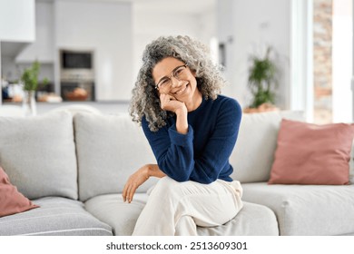 Happy 50 years old woman sitting on sofa at home looking at camera. Smiling middle aged lady posing in living room at home. Mature older female with curly gray hair relaxing on couch. Portrait. - Powered by Shutterstock