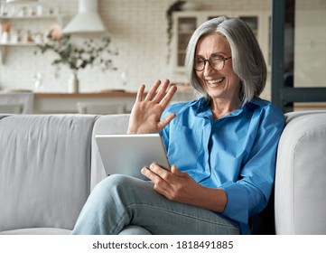 Happy 60s Older Mature Middle Aged Adult Woman Waving Hand Holding Digital Tablet Computer Video Conference Calling By Social Distance Virtual Family Online Chat Meeting Sitting On Couch At Home.