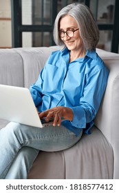 Happy 60s Older Mature Middle Aged Adult Woman Holding Laptop Using Computer Sitting On Couch At Home. Smiling Elegant Senior Grey-haired Lady Spending Time With Technology Device In Living Room.