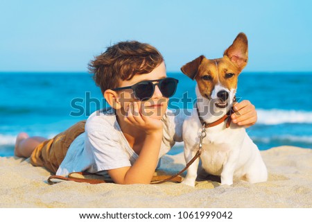 Happy 8 year old boy hugging his dog breed Jack Russell terrier at the seashore against a blue sky close up at sunset. Best friends rest and have fun on vacation, play in the sand against the sea