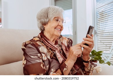 Happy 90s Older Mature Adult Woman Using Smartphone Sitting On Couch At Home. Cheerful Grandmother With Eyeglasses Surfing Internet With Her Mobile Phone. Technology Concept.