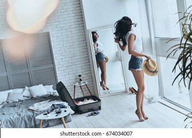 Happy About Summer Vacation. Full Length Of Beautiful Young Woman Holding Sun Hat While Looking In The Mirror At Home