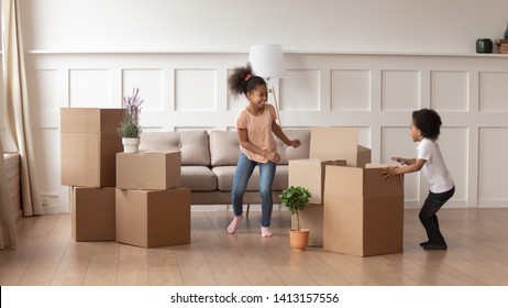 Happy Active African American Kids Playing On Moving Day, Two Excited Funny Cute Little Mixed Race Children Boy And Girl Running Around Cardboard Boxes Laughing In Living Room In New House Apartment