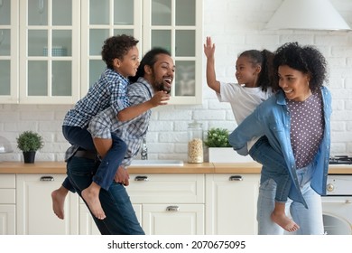 Happy Active African Parents Piggybacking Sibling Children, Running In Kitchen, Chasing Each Other Dancing. Energetic Family Couple Carrying Kids On Backs, Running, Playing Funny Games At Home