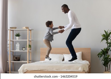 Happy Active Black Father And Cute Small Mixed Race Kid Son Holding Hand Jumping On Bed Mattress, Carefree African American Family Dad With Little Child Boy Having Fun Laughing In Bedroom Together